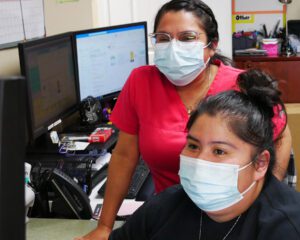 Two Hispanic clinic staff members in masks stand behind a computer looking at the screen.