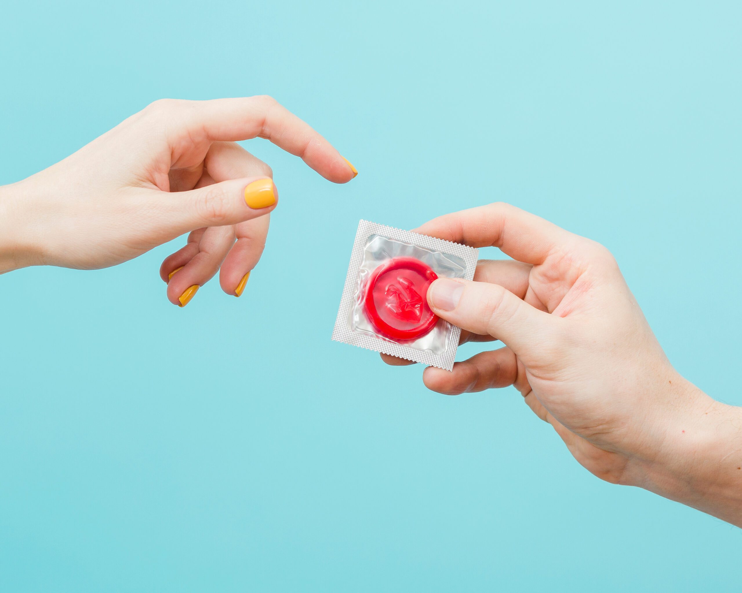 Tow hands on a blue background, one is handing the other a red condom in a clear condom wrapper.